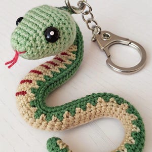 Puede incluir: Llavero de serpiente de ganchillo en tonos verdes y beige, con lengua roja y ojos negros. La serpiente está unida a un llavero y cierre plateados. El artículo hecho a mano es un accesorio caprichoso.