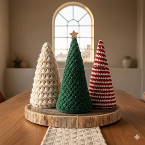 Könnte beinhalten: Drei handgefertigte gehäkelte Weihnachtsbäume in Creme, Grün und rot-weiß gestreift. Der grüne Baum hat einen Holzstern oben und silberne Perlen. Die Bäume werden auf einer Holzplatte ausgestellt, ideal für die Feiertagsdekoration.