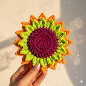 May include: A handmade, crocheted sunflower coaster. The coaster features a vibrant color palette, with a deep purple center, surrounded by layers of bright green, orange, and yellow petals. The coaster is held in a hand, showcasing the texture and detail of the yarn.