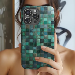 以下が含まれることがあります： 緑とティール色のモザイクタイルのデザインのスマートフォンケース。ケースは正方形のパターンで、3つのレンズを備えたカメラモジュールが付いています。ケースは人の前にかざされています。