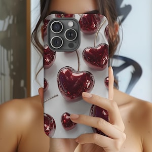 May include: A smartphone case featuring a pattern of glossy, dark red cherries with gold stems against a white background. The case has a camera cutout and is held in front of a person.