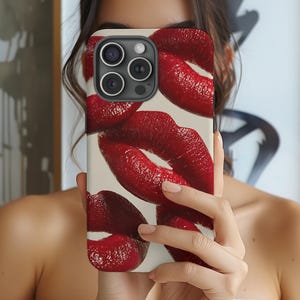 May include: A phone case featuring a design of multiple red lips against a cream-colored background. The case is held up in front of a person's face, showcasing the bold lip print.