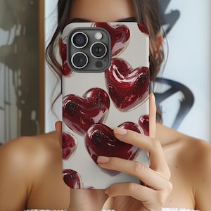 May include: A smartphone case featuring a pattern of glossy, red heart shapes against a light background. The case has a camera module with multiple lenses. The overall design is romantic and modern.