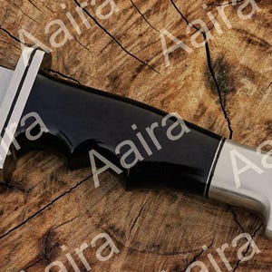 May include: Close-up of a knife with a black handle and a silver blade. The handle has a glossy finish and a curved design. The blade is partially visible, with a sharp edge. The knife is set against a wooden background.