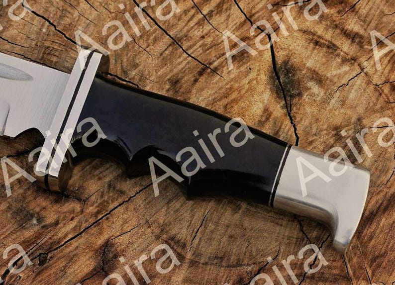 May include: Close-up of a knife with a black handle and a silver blade. The handle has a glossy finish and a curved design. The blade is partially visible, with a sharp edge. The knife is set against a wooden background.