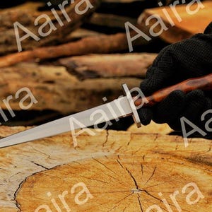 May include: A dagger with a polished steel blade and a wooden handle. The handle is a dark brown color with a silver metal accent. The dagger is held against a wooden surface.