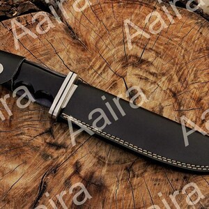 May include: A black hunting knife with a silver bolster and a black leather sheath. The knife is resting on a wooden surface. The sheath has white stitching along the edges.
