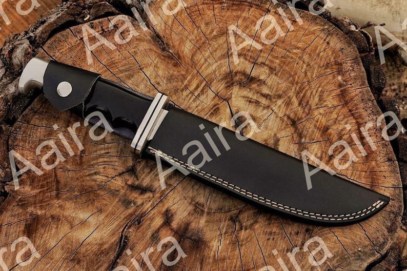 May include: A black hunting knife with a silver bolster and a black leather sheath. The knife is resting on a wooden surface. The sheath has white stitching along the edges.