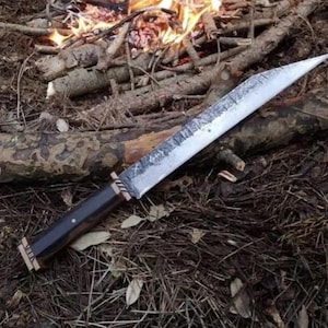 May include: A large, handmade knife with a long, textured blade and a dark wooden handle. The knife has a sharp point and is resting on a bed of dry leaves and twigs, with a campfire in the background. The knife appears to be a survival or hunting tool.