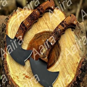 May include: Two small, curved knives with dark blades and intricately carved wooden handles. The handles feature a geometric pattern and are accented with small white dots. The knives rest on a wooden surface.