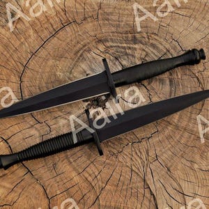 May include: Two black daggers with double-edged blades and textured handles are displayed on a wooden surface. The daggers have a crossguard and a pointed tip. The wooden surface has a natural, rustic appearance.