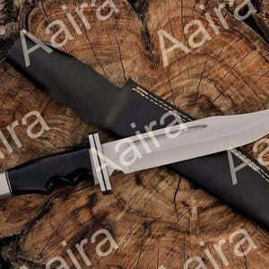 May include: A large, silver-bladed knife with a black handle and a black leather sheath. The knife has a full tang construction and a polished finish. The sheath is stitched with white thread. The knife is displayed on a wooden surface.
