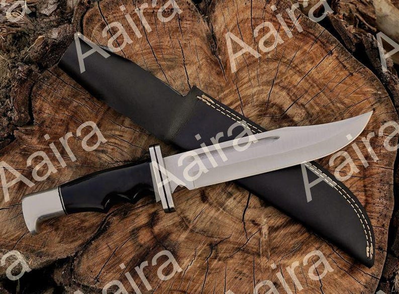 May include: A large, silver-bladed knife with a black handle and a black leather sheath. The knife has a full tang construction and a polished finish. The sheath is stitched with white thread. The knife is displayed on a wooden surface.