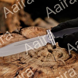 May include: A large, silver-bladed knife with a black handle and a silver bolster is held by a gloved hand. The knife rests on a textured, brown wooden surface, showcasing its sharp edge and design. The blade appears to be approximately 8 inches long.