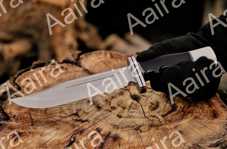 May include: A large, silver-bladed knife with a black handle and a silver bolster is held by a gloved hand. The knife rests on a textured, brown wooden surface, showcasing its sharp edge and design. The blade appears to be approximately 8 inches long.