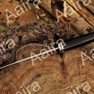 May include: A black and silver knife with a long, thin blade. The handle is black with a silver end. The knife is lying on a weathered, brown wooden surface.