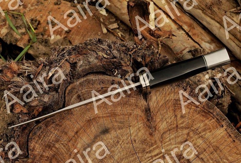 May include: A black and silver knife with a long, thin blade. The handle is black with a silver end. The knife is lying on a weathered, brown wooden surface.