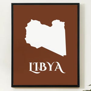 May include: Framed art print featuring a white silhouette of Libya on a brown background. The word "LIBYA" is displayed in elegant white lettering below the map. The artwork is encased in a black frame.