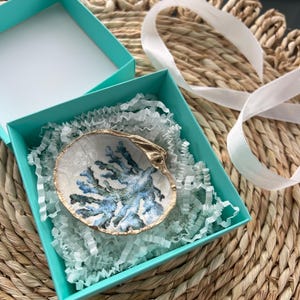 May include: A small, white seashell ring dish with a blue and green coral design. The dish is in a teal blue gift box with white tissue paper.