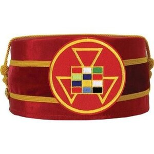May include: A red fez hat with gold trim and a circular emblem featuring a geometric design and colorful squares. The hat has a braided gold cord on each side.