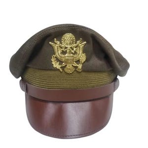 Könnte beinhalten: Eine Militärmütze mit brauner Krone, khakifarbenem Band und braunem Lederschirm. Ein goldfarbenes Emblem ist mittig auf der Vorderseite angebracht. Die Mütze hat einen Lederriemen und eine abgerundete Oberseite.