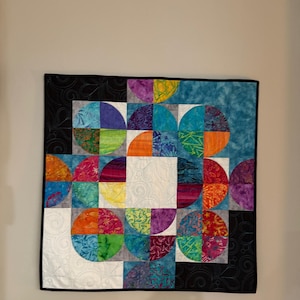 May include: A colorful, geometric quilt with a modern design. The quilt features a pattern of half-circles in various colors, including blue, orange, purple, and green, set against a black and white background. The quilt is framed with a black border.