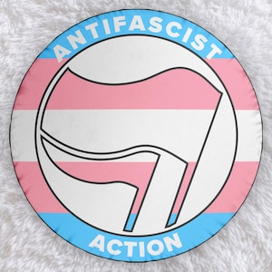 Könnte beinhalten: Ein runder, blauer und rosa Kreis mit dem Text "ANTIFASCIST ACTION" in Weiß. Der Kreis enthält eine stilisierte, schwarz-weiße Faust, die eine Flagge mit den Farben der Transgender-Pride-Flagge hält.