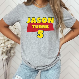 May include: A heather gray short-sleeve t-shirt featuring a graphic print with the text JASON TURNS 5. The word JASON is in yellow, the word TURNS is in yellow on a red trapezoid background, and the number 5 is in large yellow font.