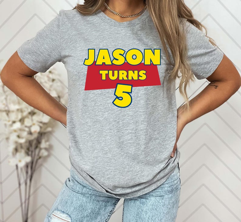 May include: A heather gray short-sleeve t-shirt featuring a graphic print with the text JASON TURNS 5. The word JASON is in yellow, the word TURNS is in yellow on a red trapezoid background, and the number 5 is in large yellow font.
