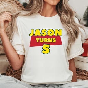 May include: A white short-sleeved t-shirt featuring a graphic print with the text JASON in yellow, TURNS in yellow on a red rectangular background, and the number 5 in yellow with a blue outline, centered on the chest of the garment.