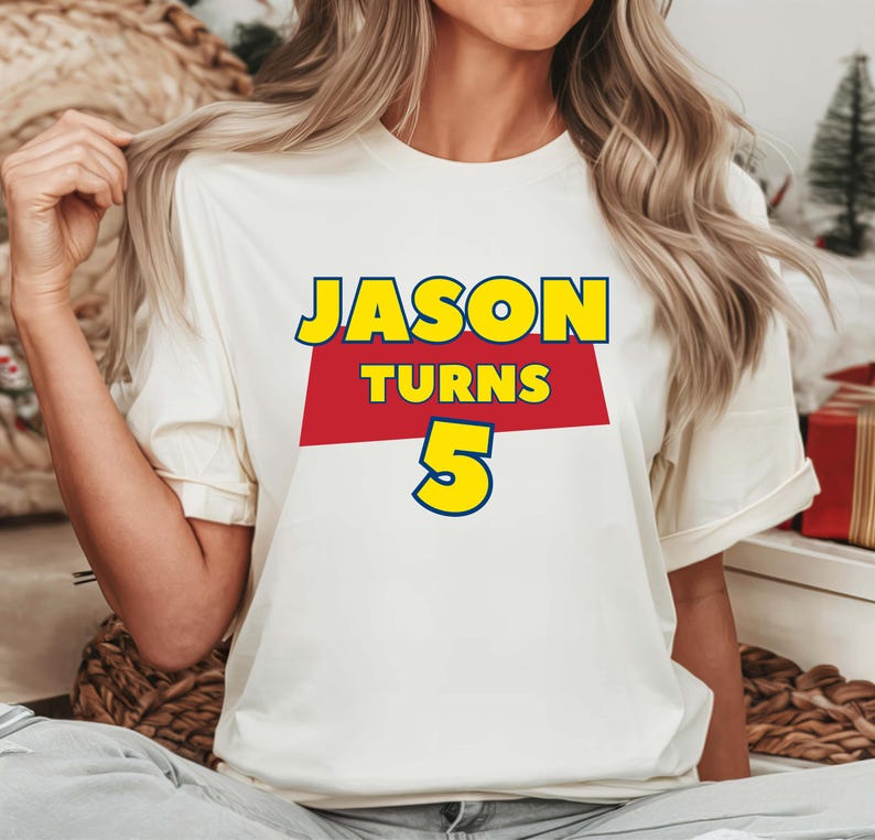 May include: A white short-sleeved t-shirt featuring a graphic print with the text JASON in yellow, TURNS in yellow on a red rectangular background, and the number 5 in yellow with a blue outline, centered on the chest of the garment.