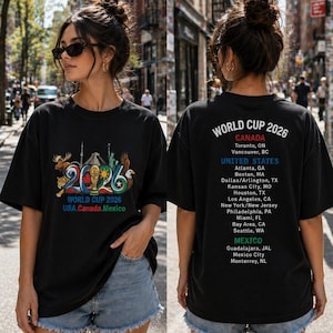 Pode incluir: Uma t-shirt preta com um gr&aacute;fico do World Cup 2026 na frente, incluindo um trof&eacute;u, s&iacute;mbolos nacionais e o texto USA, Canada, Mexico. As costas exibem o texto World Cup 2026 com uma lista das cidades-sede no Canad&aacute;, Estados Unidos e M&eacute;xico.