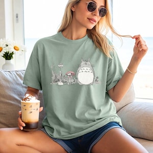 May include: Sage green t-shirt featuring a graphic of Totoro from the animated film My Neighbor Totoro. The illustration shows Totoro, a bus stop, and a character with an umbrella. The shirt is a relaxed fit.