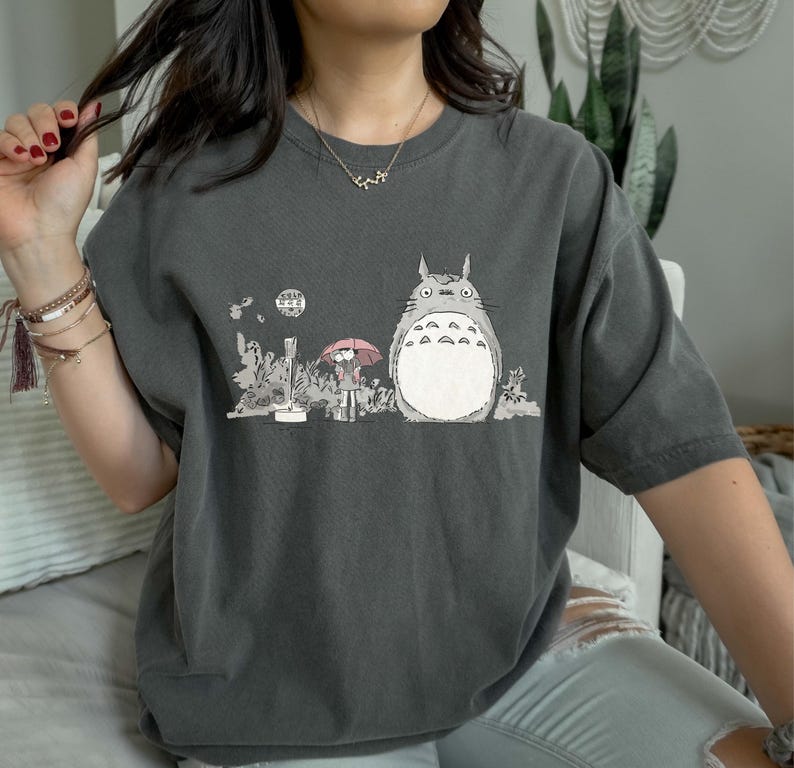 Op de afbeelding: Donkergrijs t-shirt met een illustratie van een groot, grijs personage met een witte buik, naast een persoon met een roze paraplu. Het ontwerp bevat ook een maan en bladeren.