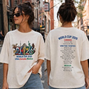 Pode incluir: Uma t-shirt em tom off-white com um gr&aacute;fico frontal contendo o texto World Cup 2026 USA.Canada.Mexico e ilustra&ccedil;&otilde;es de um alce, um jaguar e uma &aacute;guia. As costas exibem o texto World Cup 2026 com uma lista das cidades-sede.