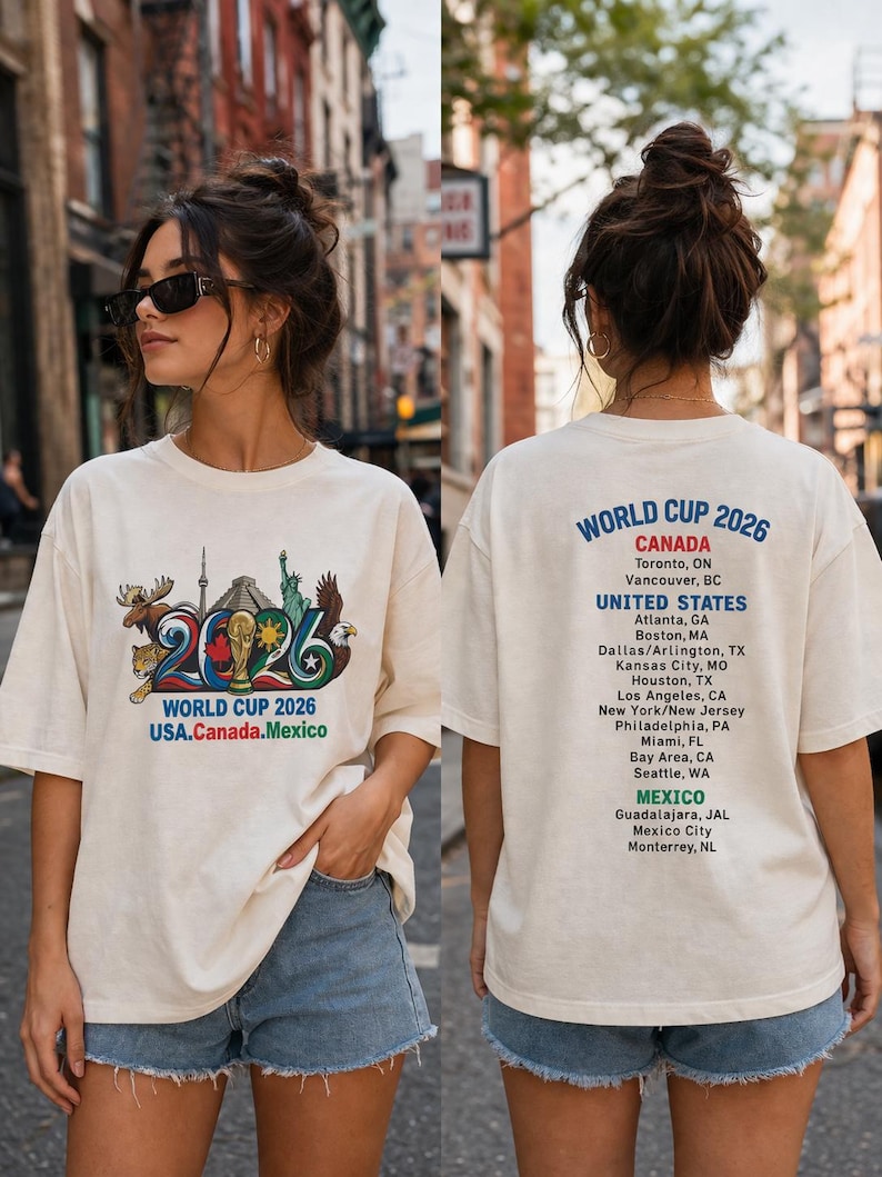 Pode incluir: Uma t-shirt em tom off-white com um gr&aacute;fico frontal contendo o texto World Cup 2026 USA.Canada.Mexico e ilustra&ccedil;&otilde;es de um alce, um jaguar e uma &aacute;guia. As costas exibem o texto World Cup 2026 com uma lista das cidades-sede.