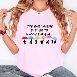 May include: Pink t-shirt with the text "The One Where They Go To Universal" and a row of colorful umbrellas with cartoon characters underneath.