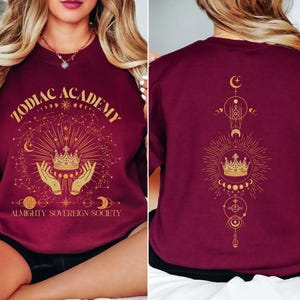 May include: Burgundy sweatshirt with a gold graphic design featuring a crown, hands, and celestial symbols. The text "Zodiac Academy Almighty Sovereign Society" is printed on the front.