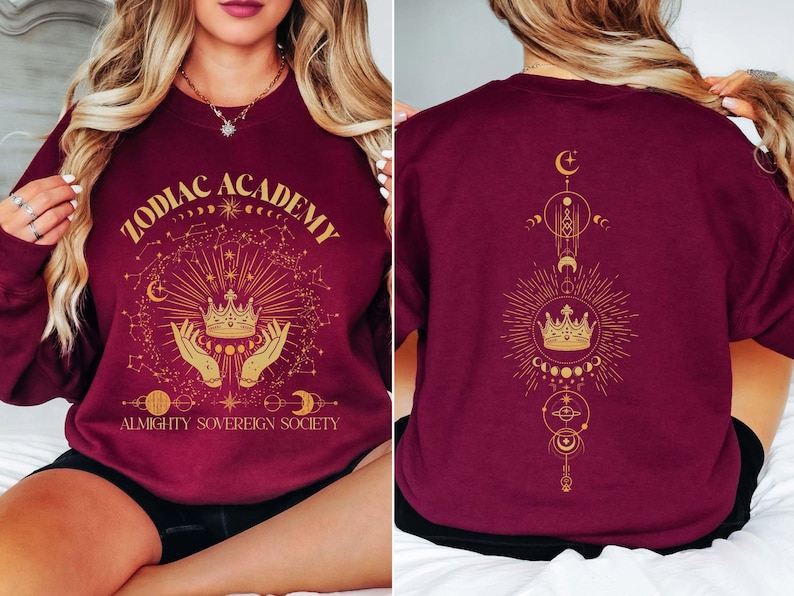 May include: Burgundy sweatshirt with a gold graphic design featuring a crown, hands, and celestial symbols. The text "Zodiac Academy Almighty Sovereign Society" is printed on the front.