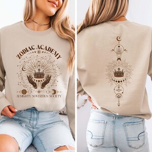 May include: A beige sweatshirt with the text "Zodiac Academy Almighty Sovereign Society" and a graphic of a crown, hands, and celestial symbols. The back of the sweatshirt features a vertical graphic of a crown, celestial symbols, and a crescent moon.