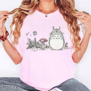 Op de afbeelding: Lichtroze T-shirt met een afbeelding van Totoro uit de animatiefilm My Neighbor Totoro. De illustratie toont Totoro naast een bushalte met een meisje met een paraplu. Het shirt is gemaakt van een zacht, comfortabel materiaal.