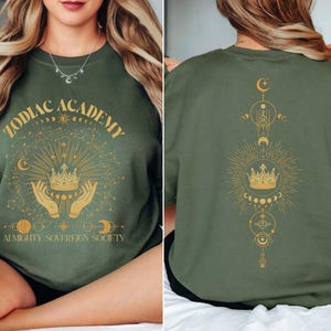 May include: Green sweatshirt with a gold graphic design featuring a crown, hands, stars, and moons. The text "Zodiac Academy Almighty Sovereign Society" is printed on the front of the sweatshirt.