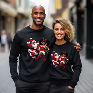 May include: Black sweatshirts featuring a cartoon Santa Claus and a woman in a Santa outfit, dancing together. The Santa figure is in a red suit with a white beard, and the woman is in a red dress with a green sash. The sweatshirts are worn by a man and a woman.