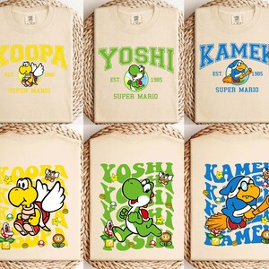 May include: Six beige t-shirts with Super Mario character designs. Each shirt features a different character name in a collegiate font: Koopa, Yoshi, and Kamek. The designs include character illustrations, the text "EST. 1985", and the words "SUPER MARIO".