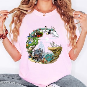 Puede incluir: Camiseta rosa claro con una colorida ilustraci&oacute;n de un drag&oacute;n y personajes de una pel&iacute;cula de animaci&oacute;n fant&aacute;stica. El gr&aacute;fico tiene forma de la letra "G". La camiseta es de material suave.