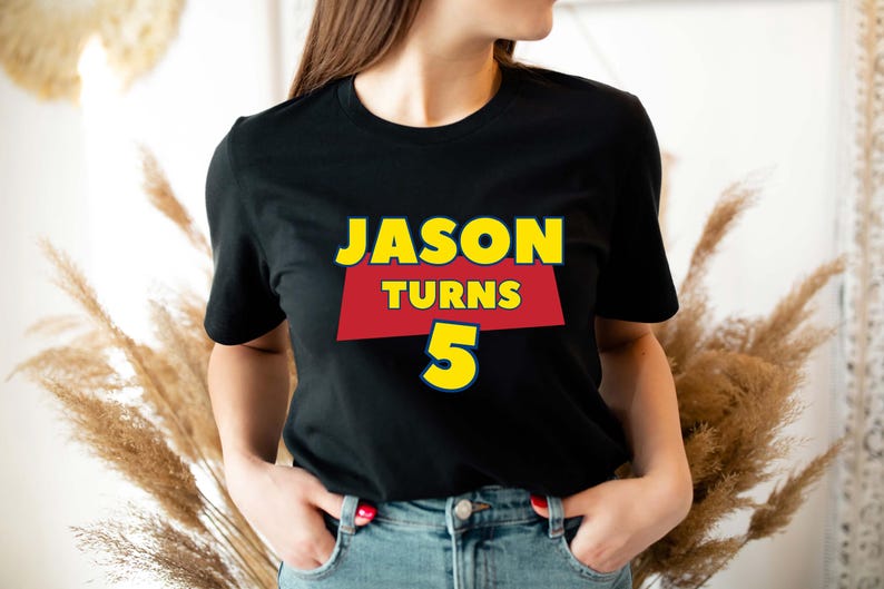 May include: A black short-sleeved t-shirt featuring a graphic design with the text JASON TURNS 5. The word JASON is in yellow, the word TURNS is in yellow on a red trapezoid background, and the number 5 is in yellow with a blue outline.