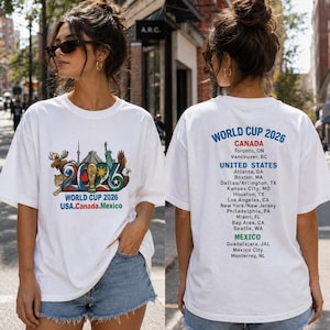 Pode incluir: Uma t-shirt branca com um gr&aacute;fico do World Cup 2026 na frente, incluindo ilustra&ccedil;&otilde;es de um alce, jaguar, &aacute;guia e monumentos. As costas exibem o texto World Cup 2026 com uma lista das cidades-sede no Canad&aacute;, Estados Unidos e M&eacute;xico.