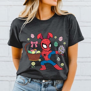 May include: Dark gray t-shirt featuring a cartoon Spider-Man character with bunny ears, holding a basket of Easter eggs and one large egg. The design includes flowers and decorative eggs, perfect for Easter.
