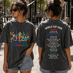 Pode incluir: Uma t-shirt cinza-carv&atilde;o com um gr&aacute;fico da Copa do Mundo 2026 na frente, ilustra&ccedil;&otilde;es coloridas de monumentos e o texto World Cup 2026 USA Canada Mexico. As costas listam as cidades-sede no Canad&aacute;, nos Estados Unidos e no M&eacute;xico.