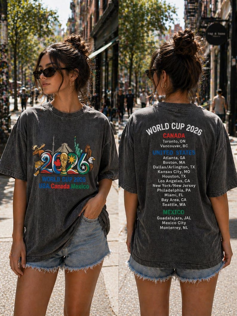 Pode incluir: Uma t-shirt cinza-carv&atilde;o com um gr&aacute;fico da Copa do Mundo 2026 na frente, ilustra&ccedil;&otilde;es coloridas de monumentos e o texto World Cup 2026 USA Canada Mexico. As costas listam as cidades-sede no Canad&aacute;, nos Estados Unidos e no M&eacute;xico.